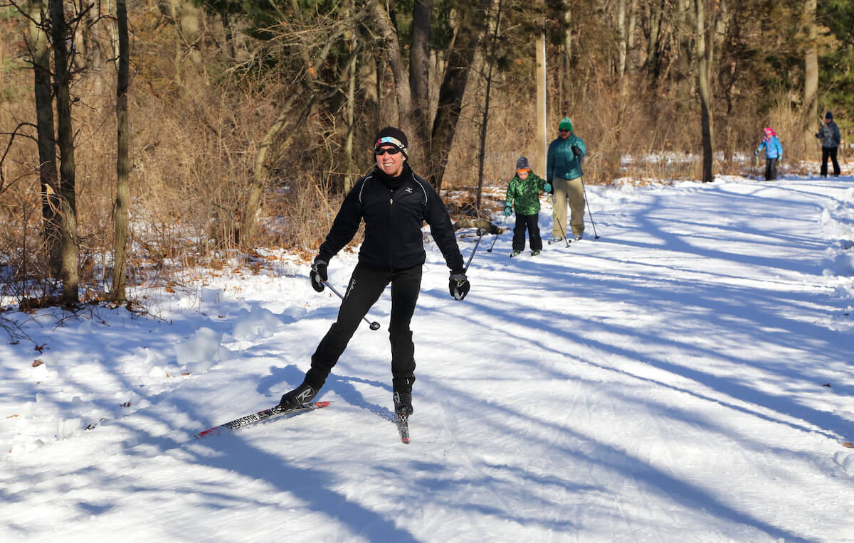 Adults and children cross-country skiing on a snowy trail through a wooded area on a sunny winter day, with trees and shadows surrounding the path.