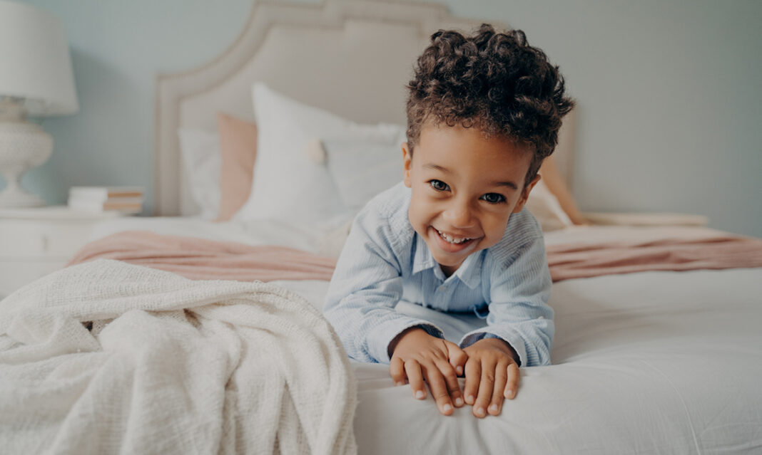 Smiling little boy in blue pajamas lying on his bed, not wanting to sleep, representing a toddler who keeps getting out of bed.