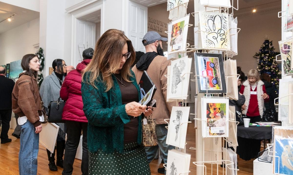 Woman browsing art cards at holiday market in gallery with people shopping and Christmas decorations in the background.