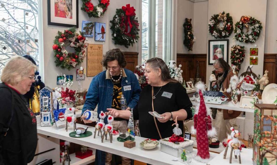 Shoppers browse handmade Christmas decorations and holiday wreaths at a festive indoor holiday market with vendors and displays.