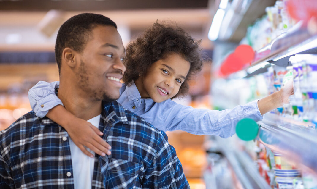 A father smiles as he shops with his young daughter, who reaches for items on the grocery shelf. The image portrays a calm and positive interaction, relevant to discussions on autism and managing aggressive behavior in public settings.