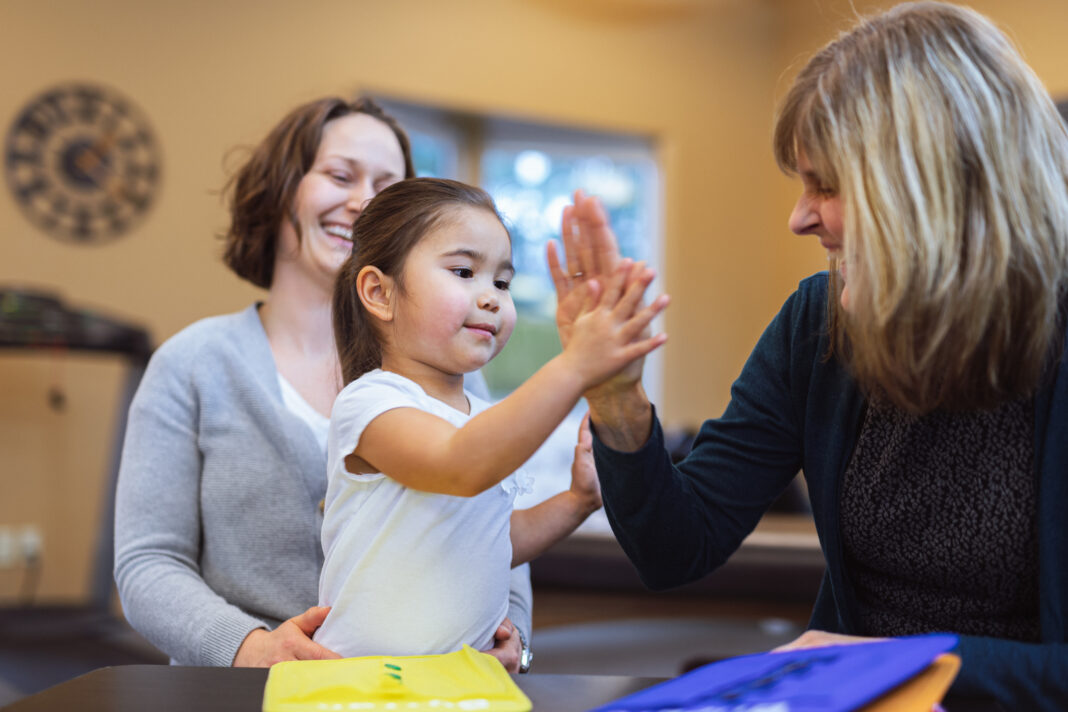 A young girl giving a high-five during a parent training ABA therapy session, with a therapist and mother supporting skill development in a positive environment.