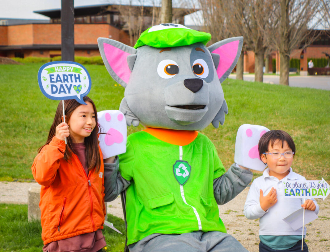 Two children pose with a recycling-themed mascot at an outdoor Earth Day celebration. The kids hold festive signs that read 