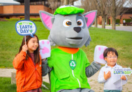 Two children pose with a recycling-themed mascot at an outdoor Earth Day celebration. The kids hold festive signs that read "Happy Earth Day" and "Go Planet, It's Your Earth Day," while the mascot wears a green hat and vest with a recycling symbol. The background features a grassy park with trees and a modern brick building.