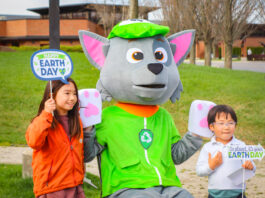 Two children pose with a recycling-themed mascot at an outdoor Earth Day celebration. The kids hold festive signs that read "Happy Earth Day" and "Go Planet, It's Your Earth Day," while the mascot wears a green hat and vest with a recycling symbol. The background features a grassy park with trees and a modern brick building.