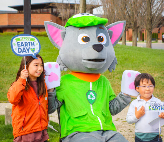 Earth Day Celebration at the Recreation Activities Center Two children pose with a recycling-themed mascot at an outdoor Earth Day celebration. The kids hold festive signs that read "Happy Earth Day" and "Go Planet, It's Your Earth Day," while the mascot wears a green hat and vest with a recycling symbol. The background features a grassy park with trees and a modern brick building.