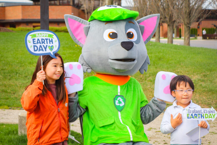 Two children pose with a recycling-themed mascot at an outdoor Earth Day celebration. The kids hold festive signs that read "Happy Earth Day" and "Go Planet, It's Your Earth Day," while the mascot wears a green hat and vest with a recycling symbol. The background features a grassy park with trees and a modern brick building.