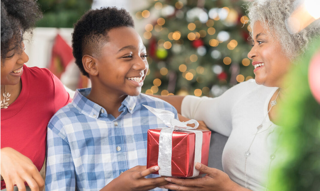 A happy boy receiving a wrapped holiday gift from family in a cozy setting with a decorated Christmas tree, showcasing the best holiday gift for metro Detroit kids.