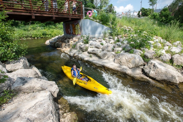 A man kayaking down a peaceful river under a bridge in Ann Arbor, a family-friendly spot for outdoor water activities.