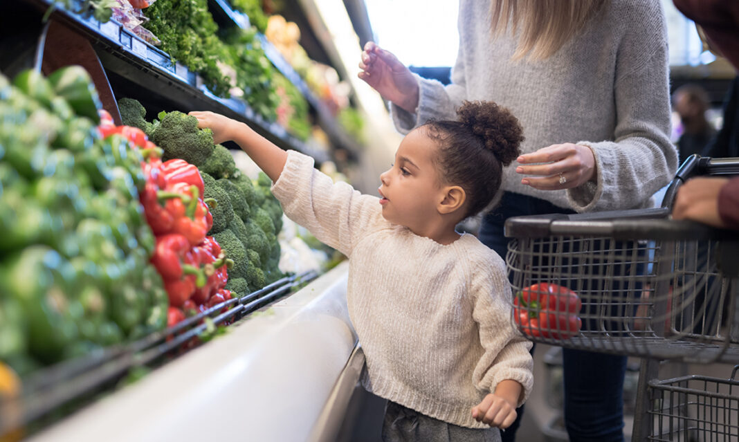 A young child reaching for fresh organic produce in a grocery store while shopping with a parent, reflecting the question 