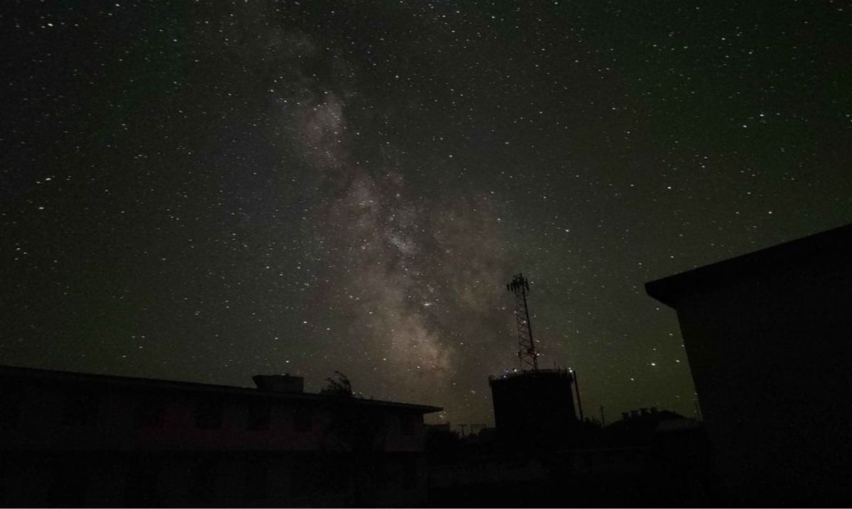 A breathtaking view of the Milky Way stretching across a dark, starry sky, with silhouettes of buildings and a communication tower in the foreground.