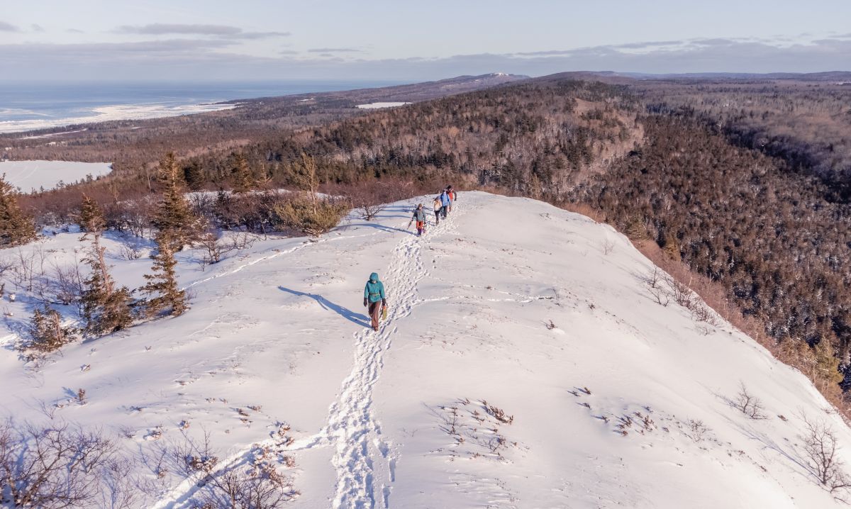 A group of hikers walking along a snow-covered ridge on a winter day in Keweenaw, surrounded by scenic forested hills and distant views of frozen lakes.