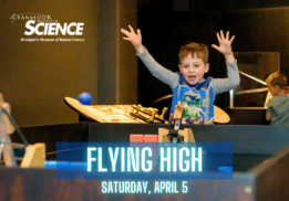 A young boy excitedly raises his hands while participating in an interactive science exhibit at the Cranbrook Institute of Science. The event, titled "Flying High," is scheduled for Saturday, April 5, and features hands-on learning experiences related to flight and physics. The background includes other children engaging with the exhibits in a museum setting.