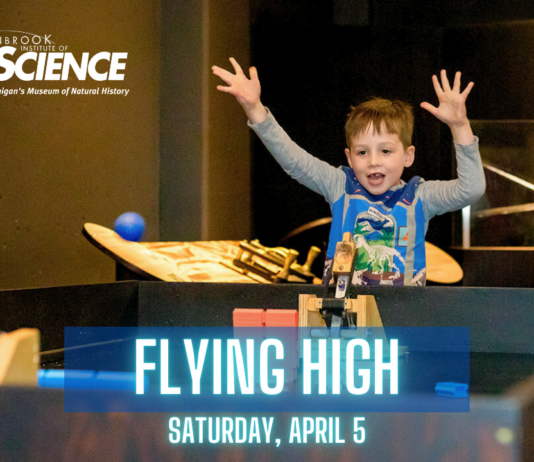 A young boy excitedly raises his hands while participating in an interactive science exhibit at the Cranbrook Institute of Science. The event, titled "Flying High," is scheduled for Saturday, April 5, and features hands-on learning experiences related to flight and physics. The background includes other children engaging with the exhibits in a museum setting.