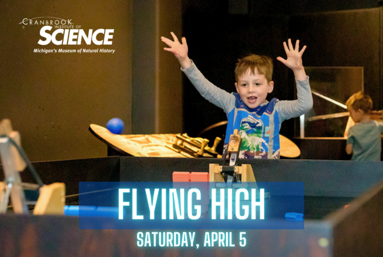 A young boy excitedly raises his hands while participating in an interactive science exhibit at the Cranbrook Institute of Science. The event, titled "Flying High," is scheduled for Saturday, April 5, and features hands-on learning experiences related to flight and physics. The background includes other children engaging with the exhibits in a museum setting.