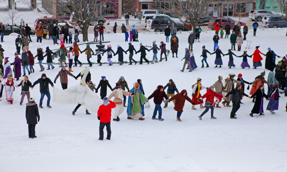 Participants holding hands in a large circle on a snowy field during the Heikinpäivä Festival, celebrating Finnish heritage with traditional winter festivities and costumes.
