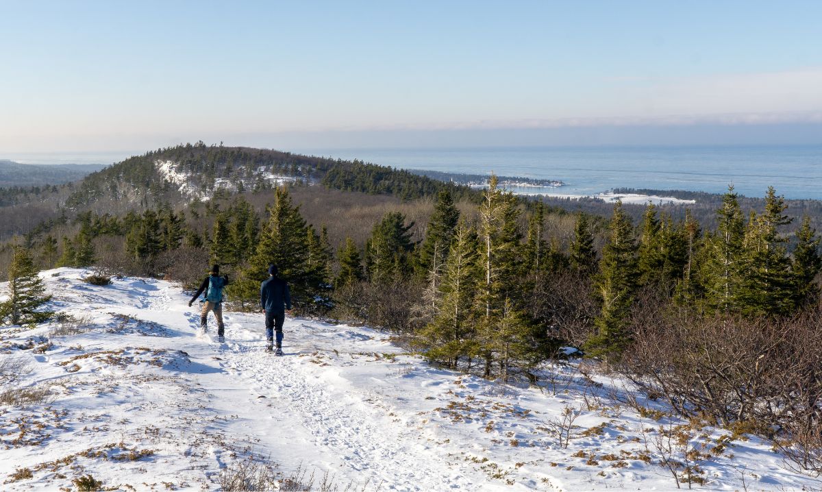 Two hikers walking along a snow-covered trail on the Keweenaw Peninsula, surrounded by evergreen trees and offering stunning views of Lake Superior and rolling hills.
