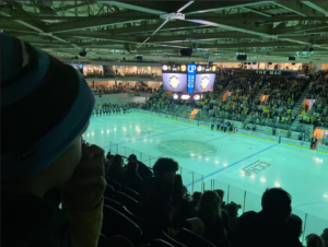 A packed crowd watching a Michigan Tech hockey game at the MacInnes Ice Arena, with the rink illuminated and the scoreboard displaying team details.