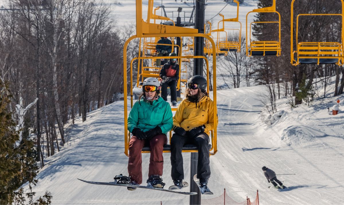 Two snowboarders riding a bright yellow ski lift at Mont Ripley, enjoying a sunny winter day with snowy slopes and trees in the background.
