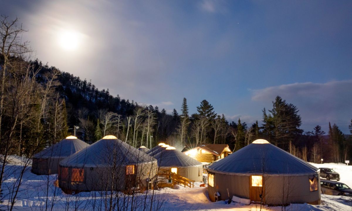 A serene nighttime view of illuminated yurts nestled in the snowy forest at Mount Bohemia, showcasing unique winter accommodations under a glowing full moon.