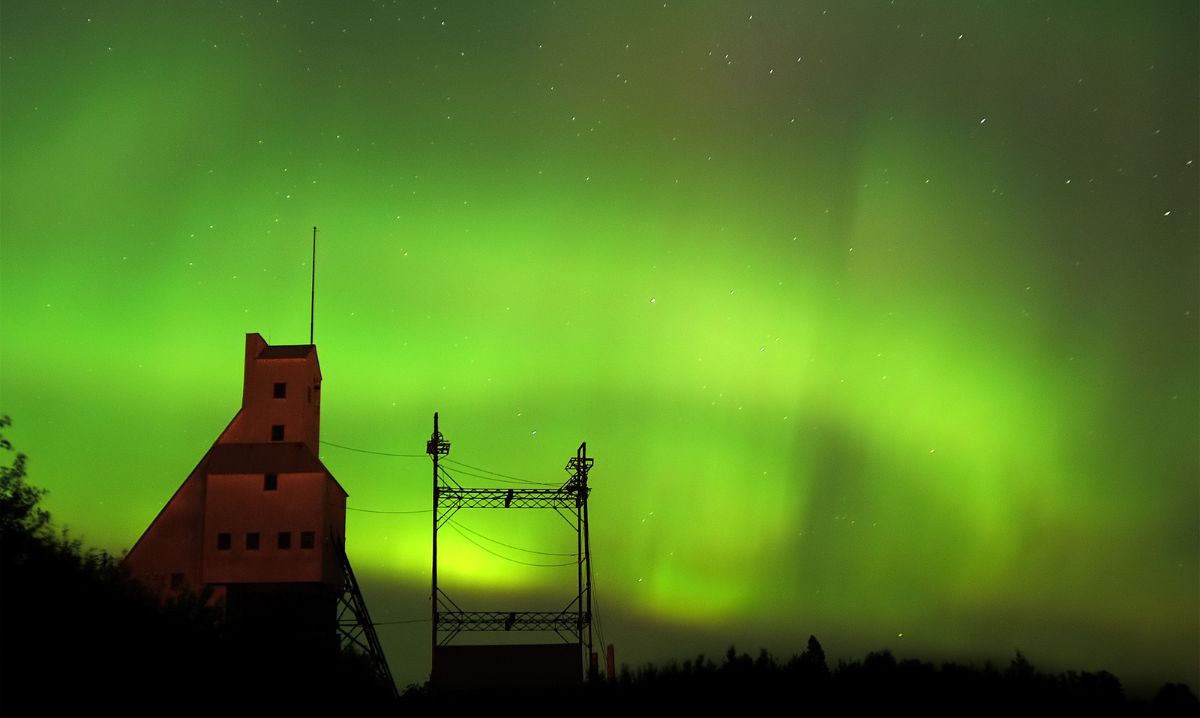 A vivid display of green Northern Lights illuminating the night sky above the Quincy Mine in Hancock, Michigan, with the mine’s silhouette creating a dramatic foreground.