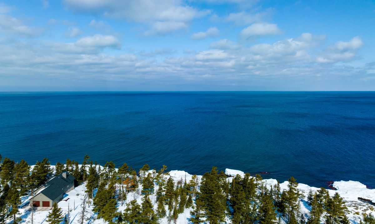 A stunning view of the winter shoreline of Lake Superior along the Keweenaw Peninsula, featuring evergreen trees, snow-covered ground, and a bright blue sky with scattered clouds.