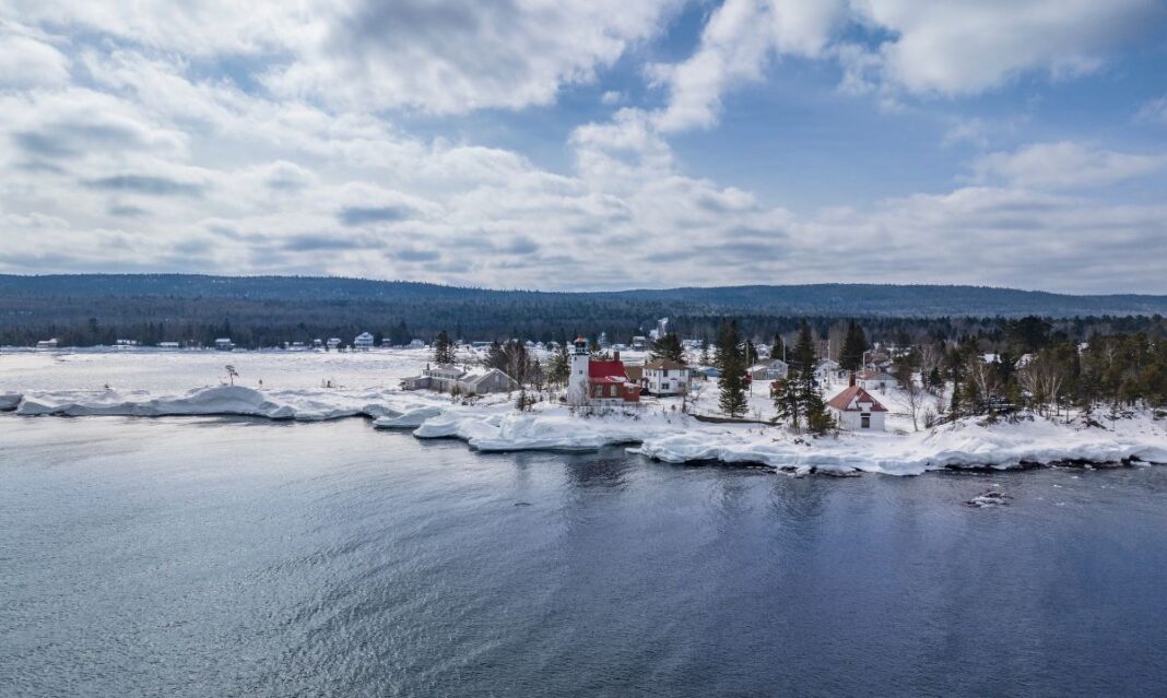 A scenic winter view of the Keweenaw Peninsula shoreline featuring a red-roofed lighthouse surrounded by snow and ice, with calm water and a wooded landscape in the background.