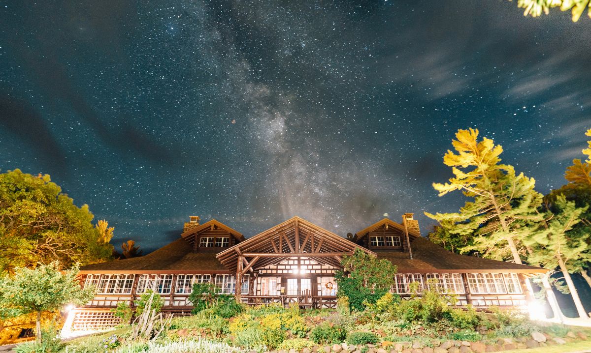 A stunning starry night with the Milky Way above a beautifully lit lodge in Keweenaw, surrounded by lush greenery and tall trees.