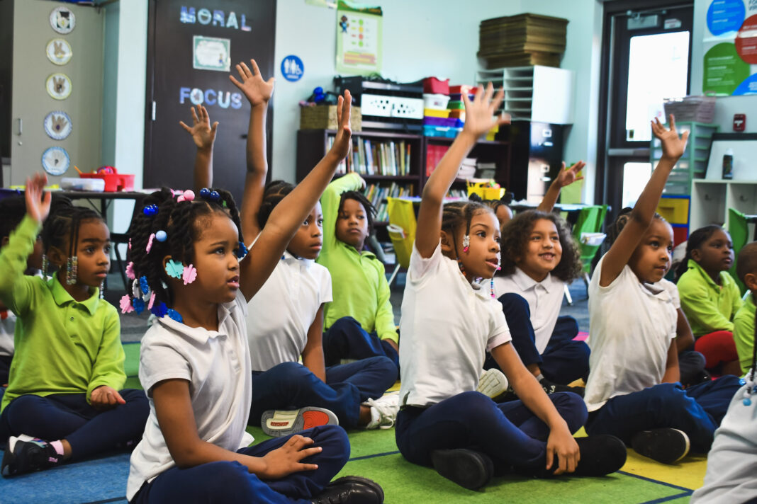 Young students sitting on a classroom rug, eagerly raising their hands to answer a question, representing engaged learning at Pembroke Academy, a National Blue Ribbon School.