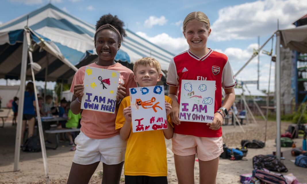 Three kids proudly holding up their artwork with the message 'I Am My Own Hero' at Camp Mirage in Troy, highlighting creative and empowering summer camp activities.