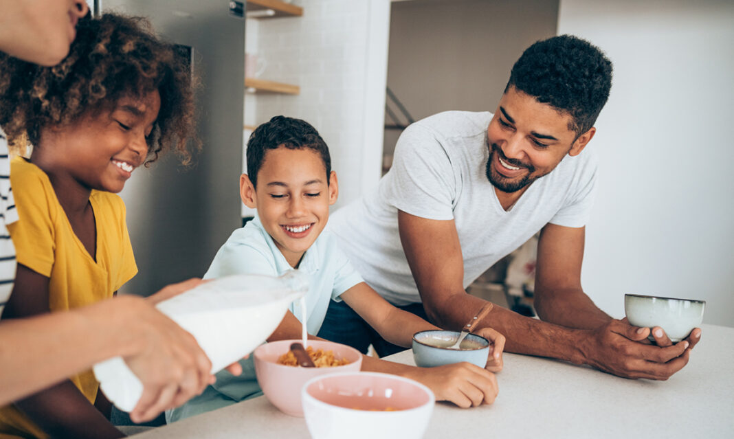 A happy family gathered in the kitchen, enjoying breakfast together. A mother is pouring milk into a bowl of cereal for her smiling daughter, with the father and son sharing a laugh nearby. This image highlights family bonding over cereal and milk recipes.