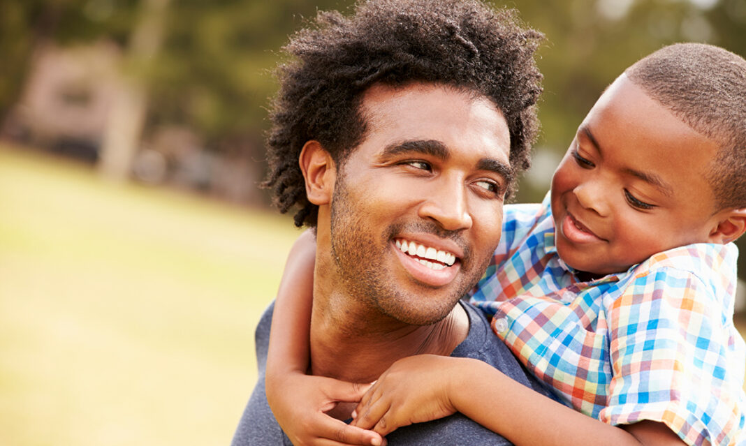 A smiling father giving his son a piggyback ride in a park. The father and son share a joyful moment outdoors, illustrating healthy coping strategies through quality family bonding and outdoor activities.