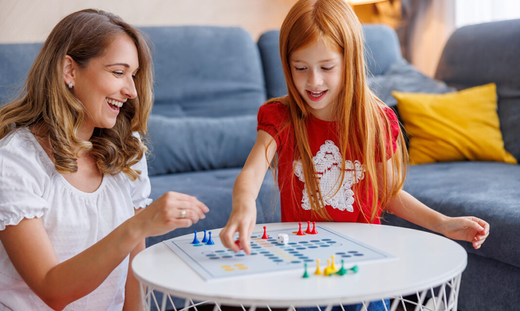A smiling mother and her young daughter playing a colorful board game together in a cozy living room, representing quality time during winter break