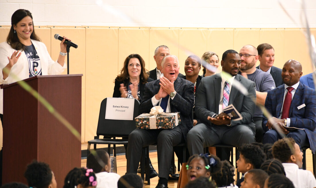 Pembroke Academy principal speaking at a National Blue Ribbon School ceremony, while community leaders and staff applaud and students watch from the audience.