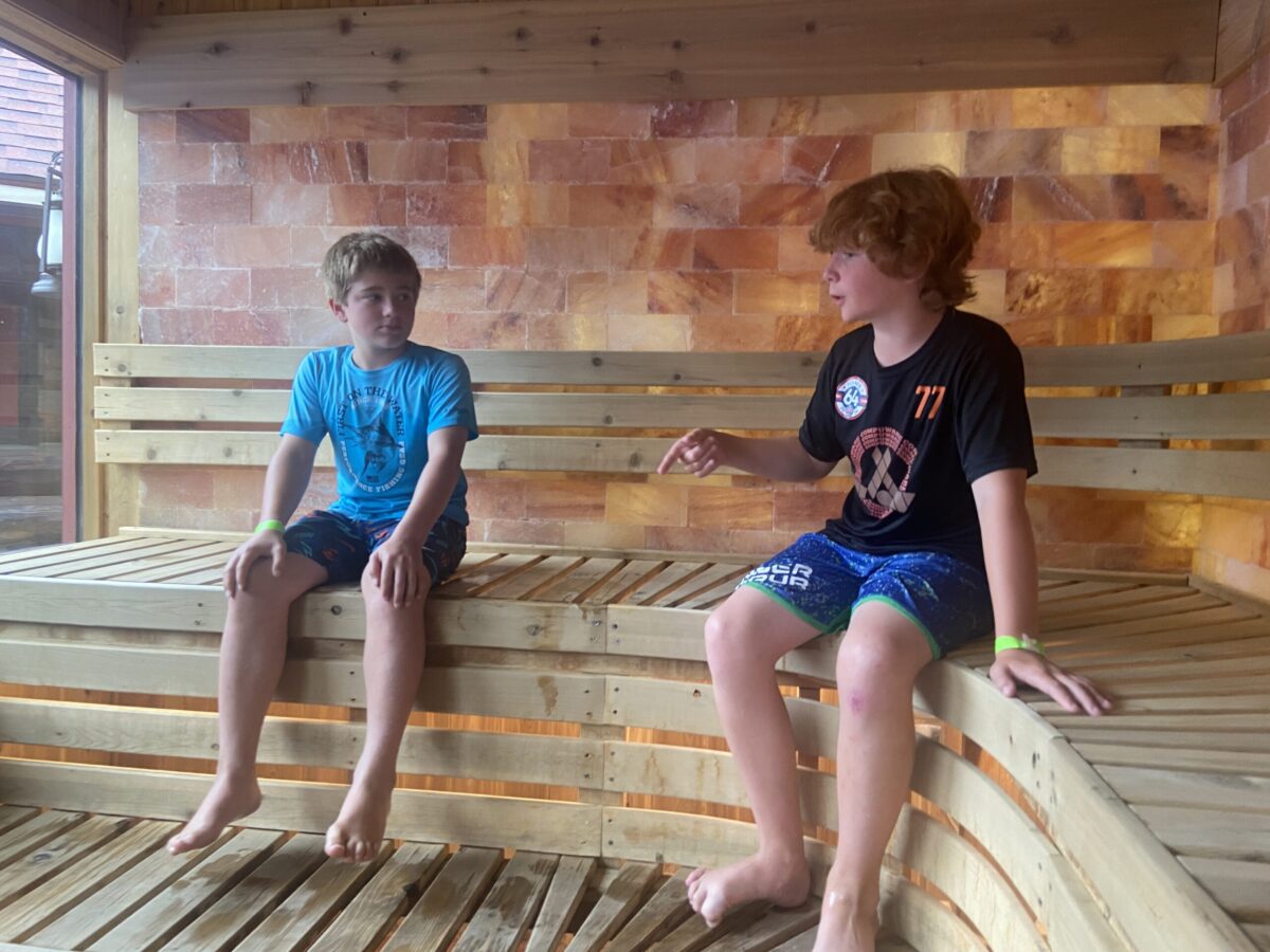 Two boys sitting and chatting in a wooden sauna with warm Himalayan salt brick walls, enjoying a relaxing and casual moment.