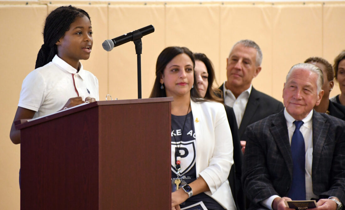 A young student speaking confidently at a podium during a National Blue Ribbon School ceremony, with Pembroke Academy school leaders and community members seated in the background.