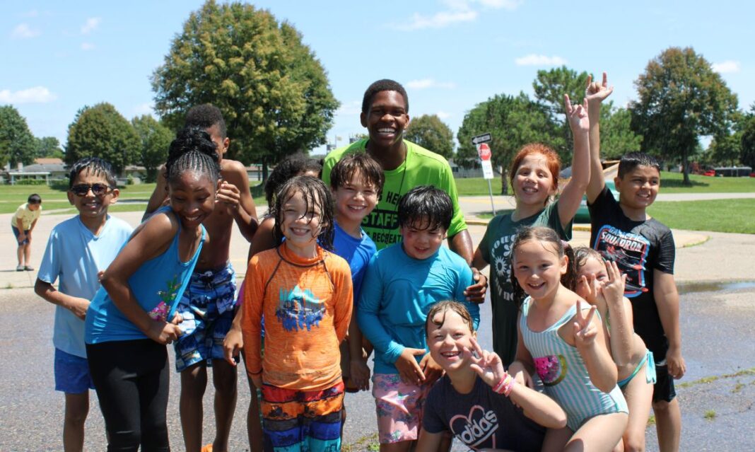 A group of smiling kids and a camp staff member enjoy a fun water play day at Summer Playground in Sterling Heights.