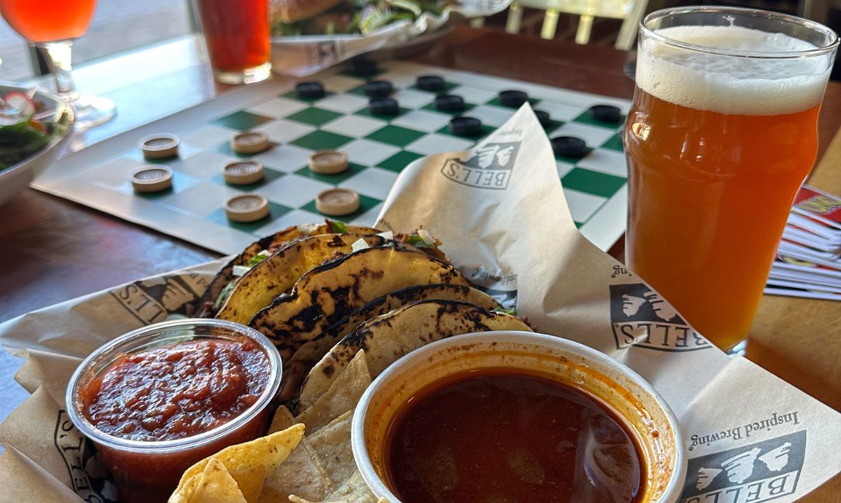 A plate of birria tacos with salsa and consommé, paired with a craft beer at Bell’s Eccentric Café in Kalamazoo, with a checkers board adding to the laid-back dining atmosphere.