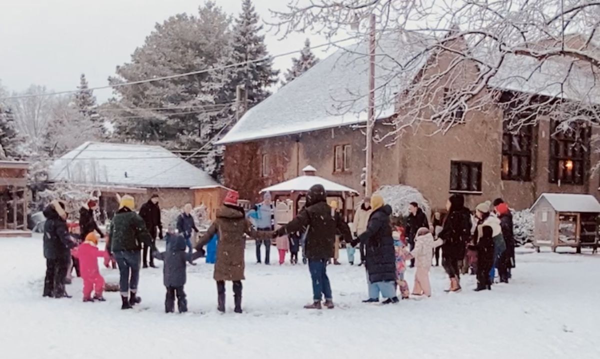 Students, teachers, and parents at Detroit Waldorf School gather outdoors in a snowy courtyard, holding hands in a community circle during a winter celebration.