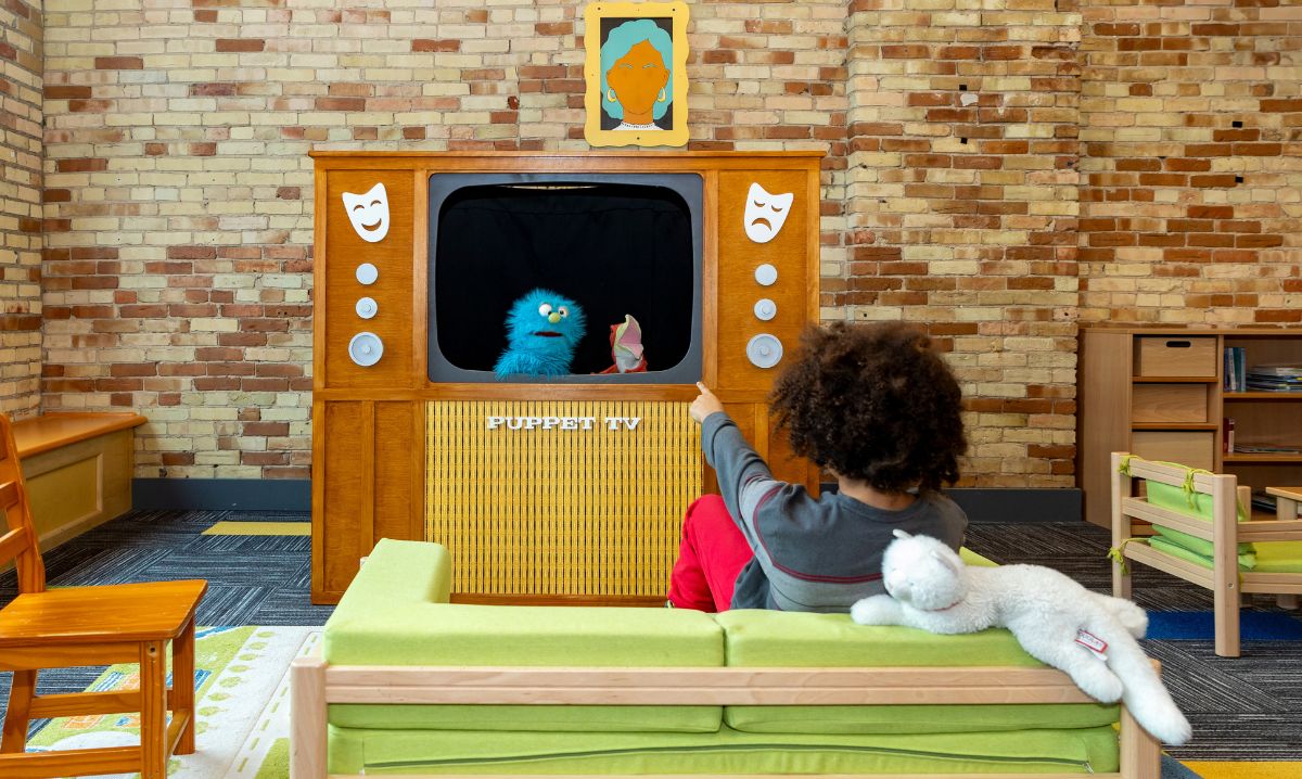 A child watches a puppet show at the Grand Rapids Children’s Museum, sitting on a small green couch with a stuffed animal in a cozy play area featuring a retro-style puppet theater.