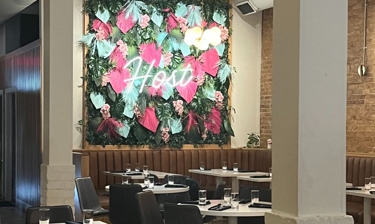 The interior of Host restaurant in Utica with a vibrant floral wall featuring pink and teal leaves, a neon sign reading 'Host,' and modern dining tables set for guests.