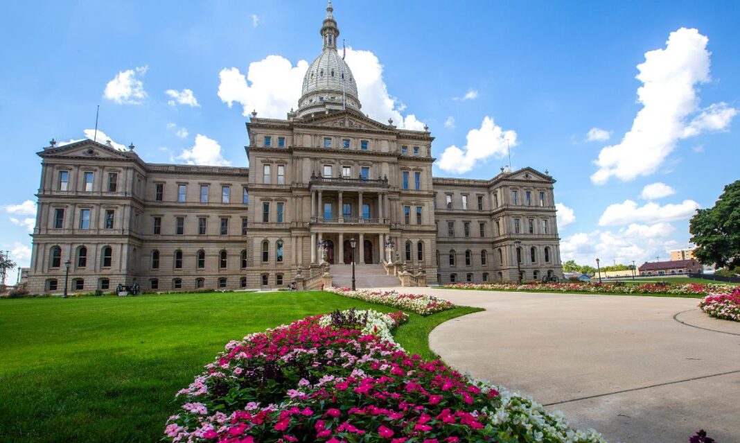 The Michigan State Capitol building in Lansing, surrounded by vibrant flower beds and a well-manicured lawn under a bright blue sky.
