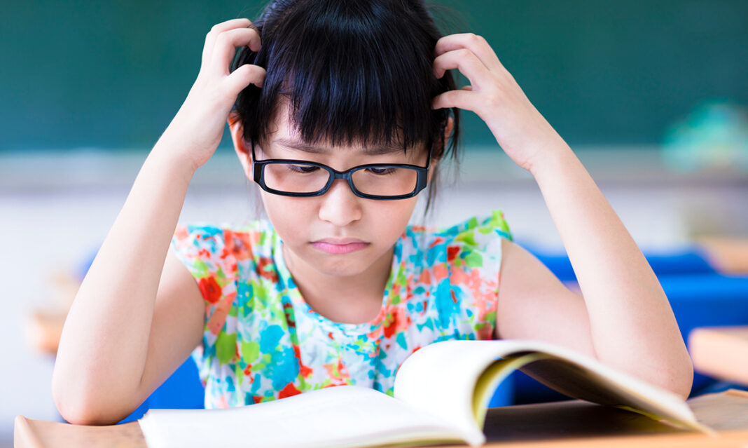 A young girl wearing glasses looks frustrated while reading a book in a classroom, holding her head in concentration.