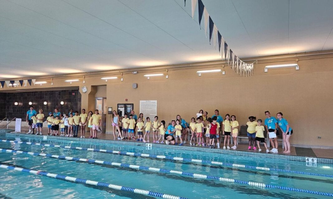 A group of kids and camp counselors in yellow and blue shirts stand poolside at Life Time Shelby Kids Camp, ready for a fun swimming session indoors.