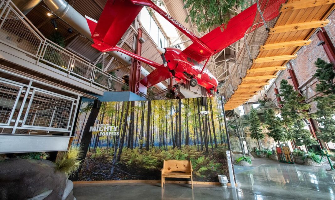 A red airplane suspended from the ceiling at the Michigan DNR Outdoor Adventure Center, with a forest-themed exhibit and a wooden suspension bridge.