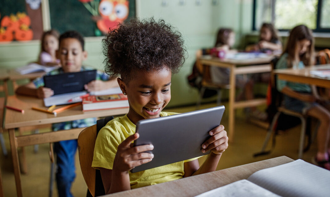 A student in a classroom using a tablet for online learning, surrounded by other children engaged in educational activities.