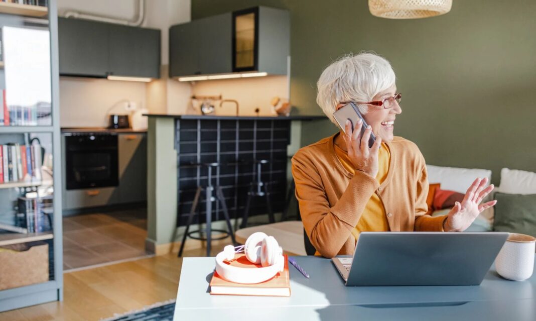 A senior woman smiling while on a phone call at home with a laptop, showcasing practical bill negotiating tips for phone calls.