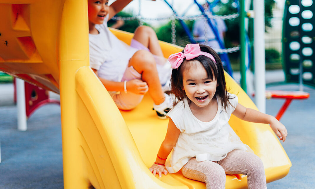 Two young children enjoying a sunny day at the playground, sliding down a bright yellow slide. The girl in the foreground, wearing a pink bow, is smiling joyfully while having fun outdoors.