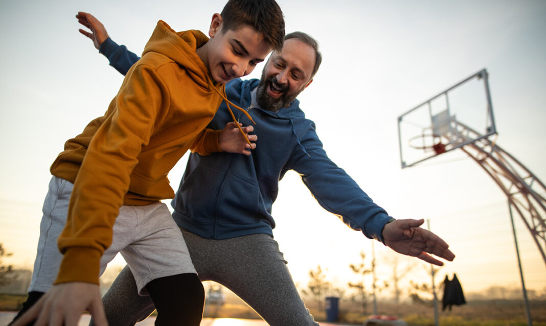 A father and son playing basketball together outdoors at sunset, sharing a joyful moment. This image represents the importance of bonding and support for siblings of children with autism through active and engaging family time.
