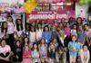 A group of smiling children and camp counselors pose at a summer camp in Rochester Hills, surrounded by colorful decorations, balloons, and fun-themed merchandise.
